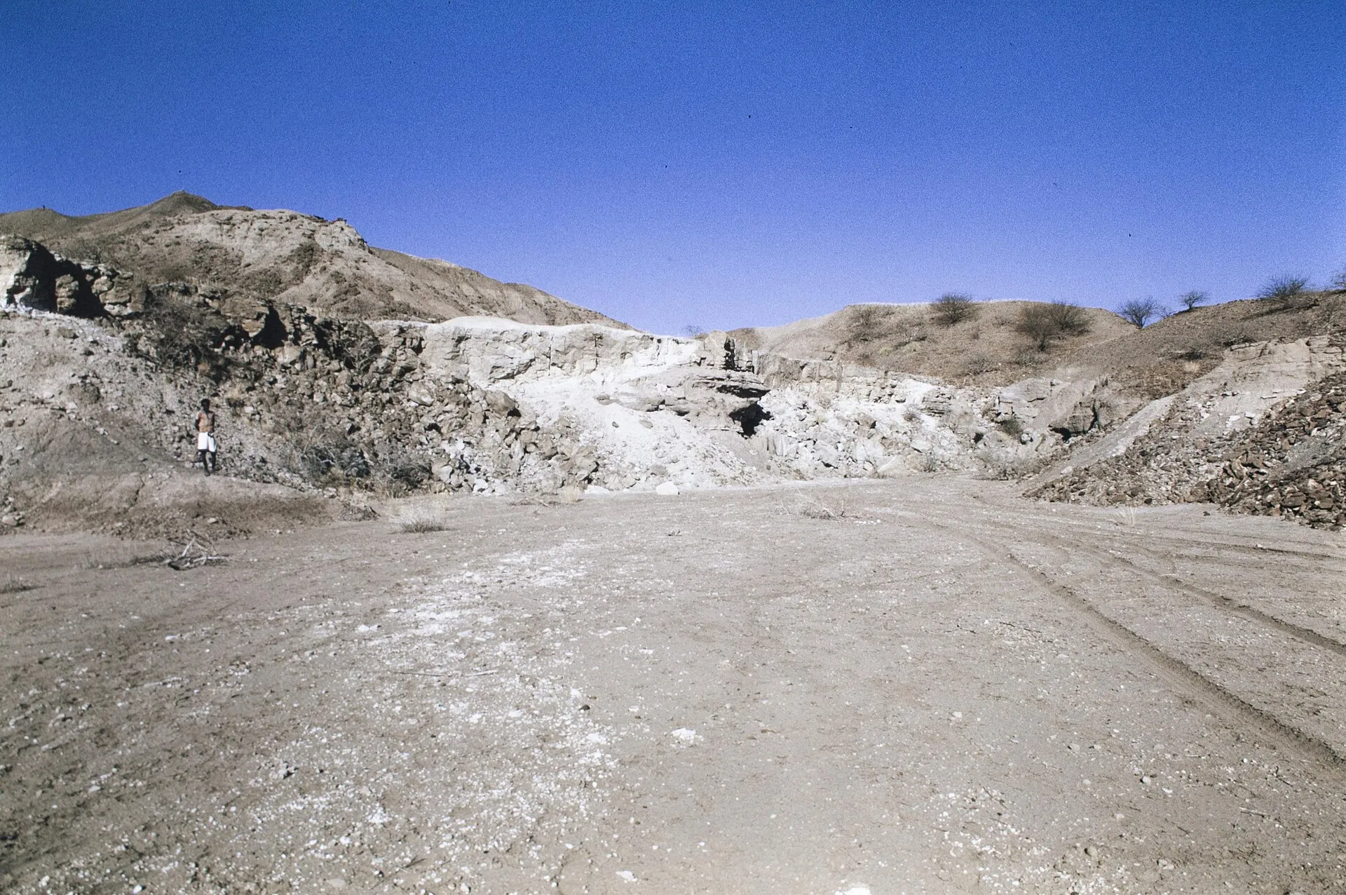 A pale, eroded hillside in the Hadar region of Ethiopia under a blue sky, with a lone figure standing at the left